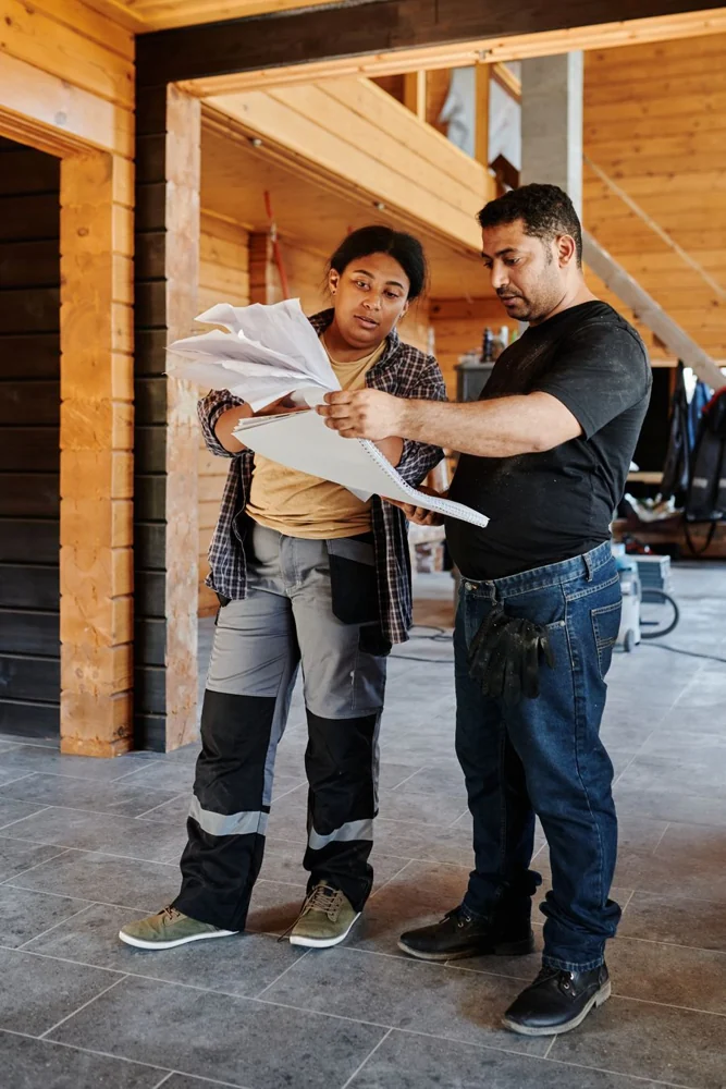 Two contractors reviewing building plans inside a timber-framed house.
